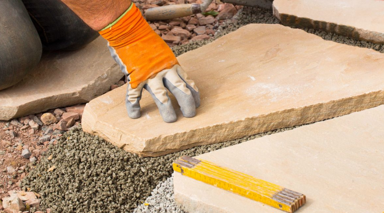 GaLaBau Hero 1 GaLaBauer bei der Arbeit kniend auf einem Naturstein mit Sicherheitshandschuhen und einem Metermaß